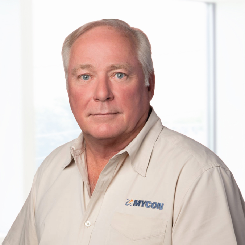 A man with gray hair wearing a beige button-up shirt with a "MYCON" logo stands in front of a bright, blurry background.