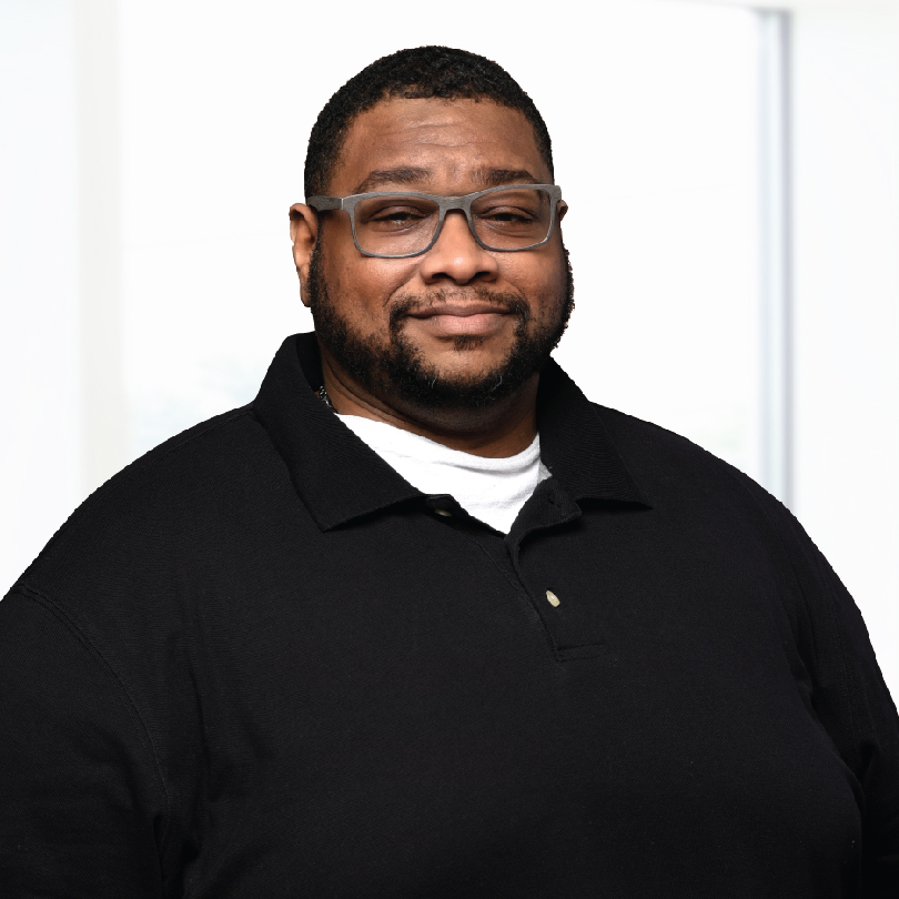 A man with short hair, beard, and glasses is wearing a black polo shirt and smiling slightly. The background is bright and blurred.
