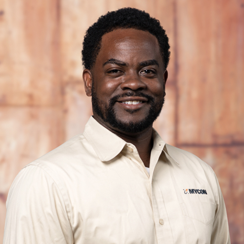 A black man in a tan shirt smiling in front of a wooden wall.