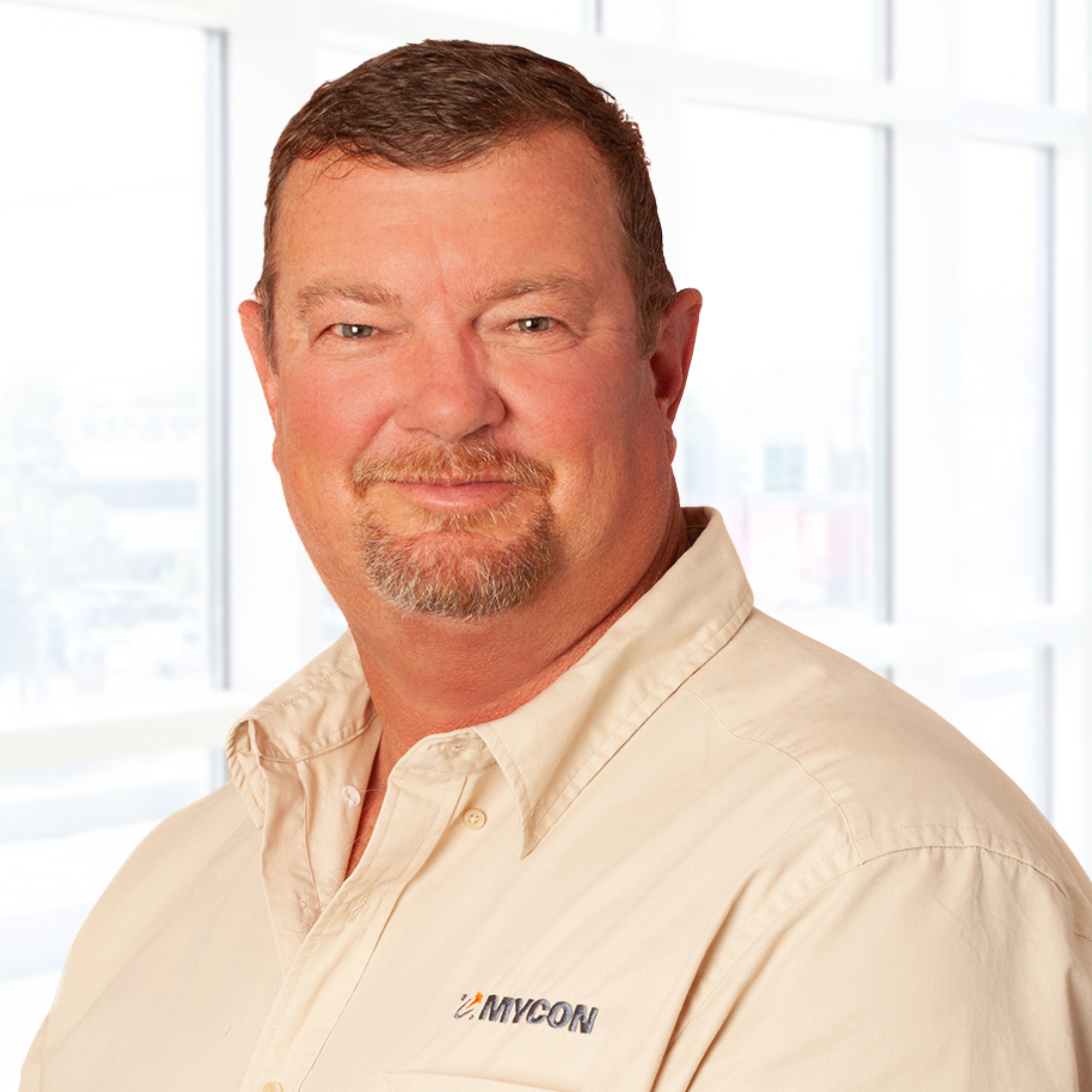 Man in a beige shirt with "MYCON" logo, standing in front of a bright window background.