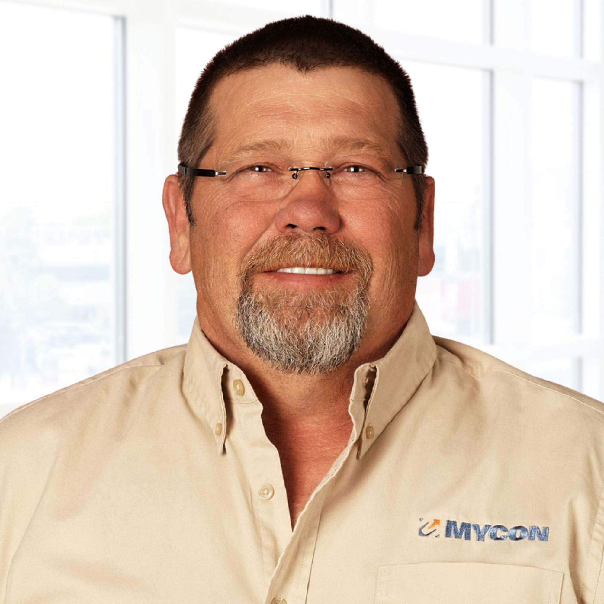Man wearing glasses and a beige shirt with a MYCON logo, posing in front of a window.