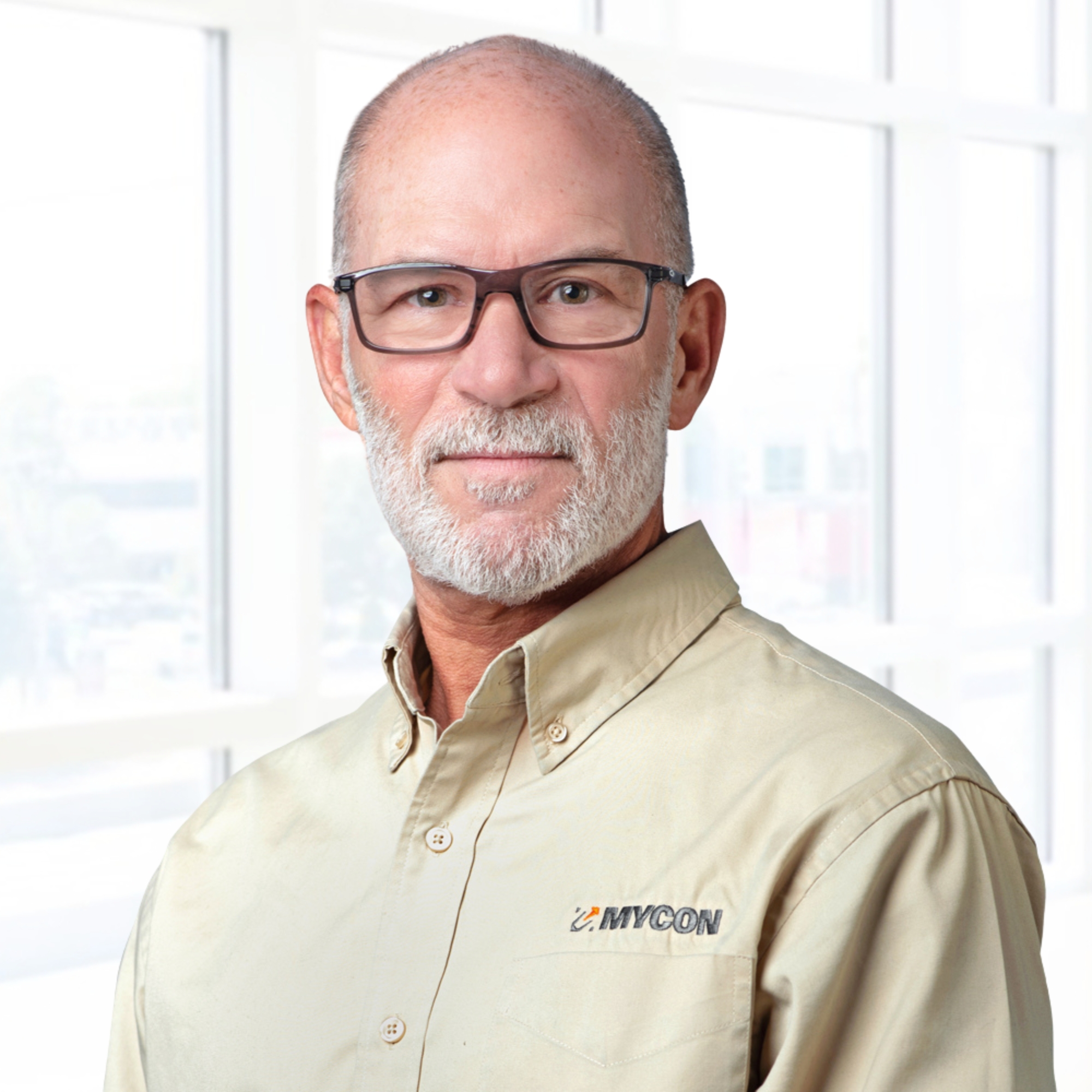 A man with glasses and a beard wearing a beige shirt with a MYCON logo, standing indoors with a bright window backdrop.