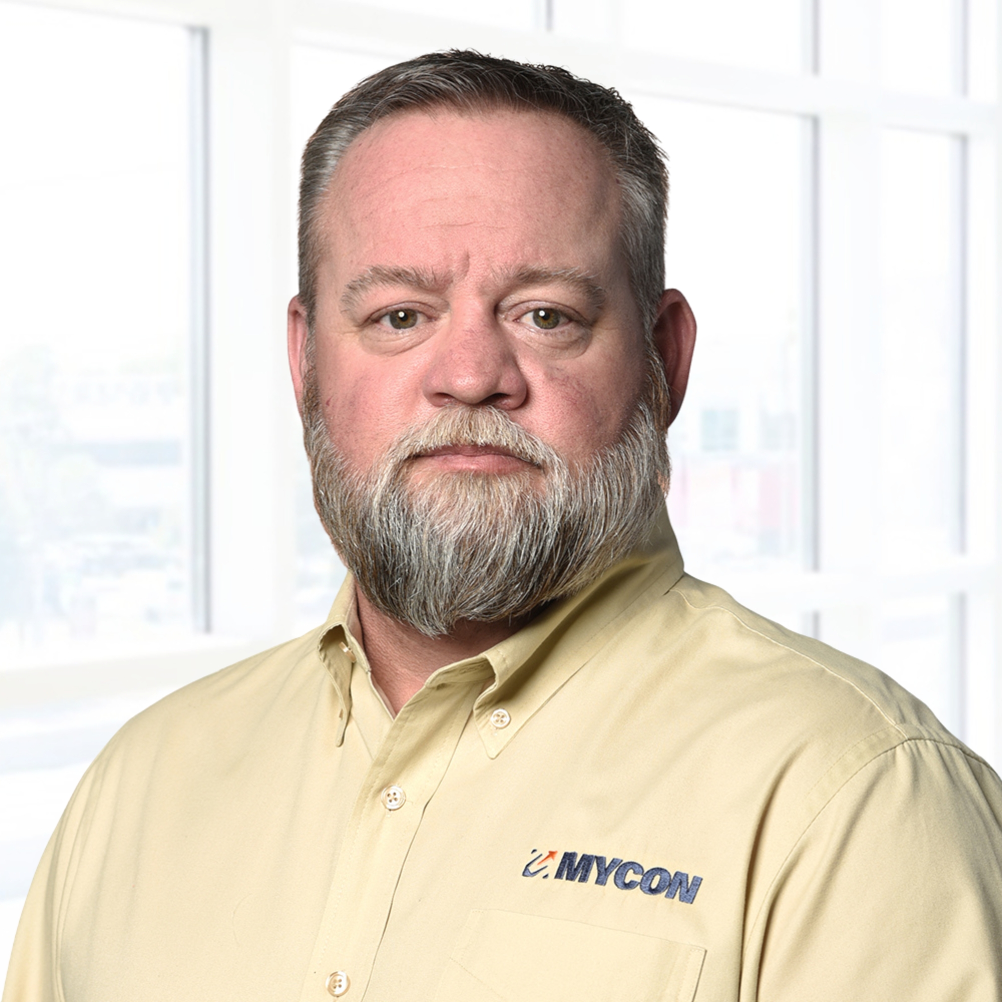 A person with a beard wearing a yellow MYCON shirt stands in front of a bright window.