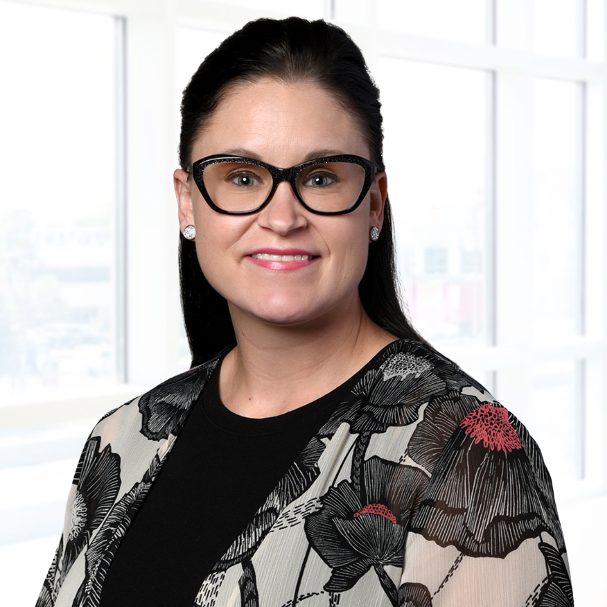 A woman with glasses and black hair smiles in a professional setting, wearing a floral-patterned jacket over a black top.