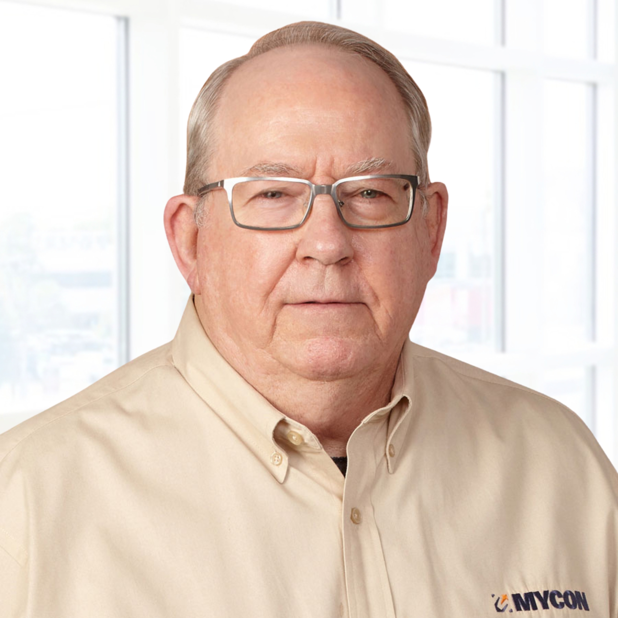 A person with gray hair and glasses, wearing a beige button-up shirt with a company logo, standing in front of a window.