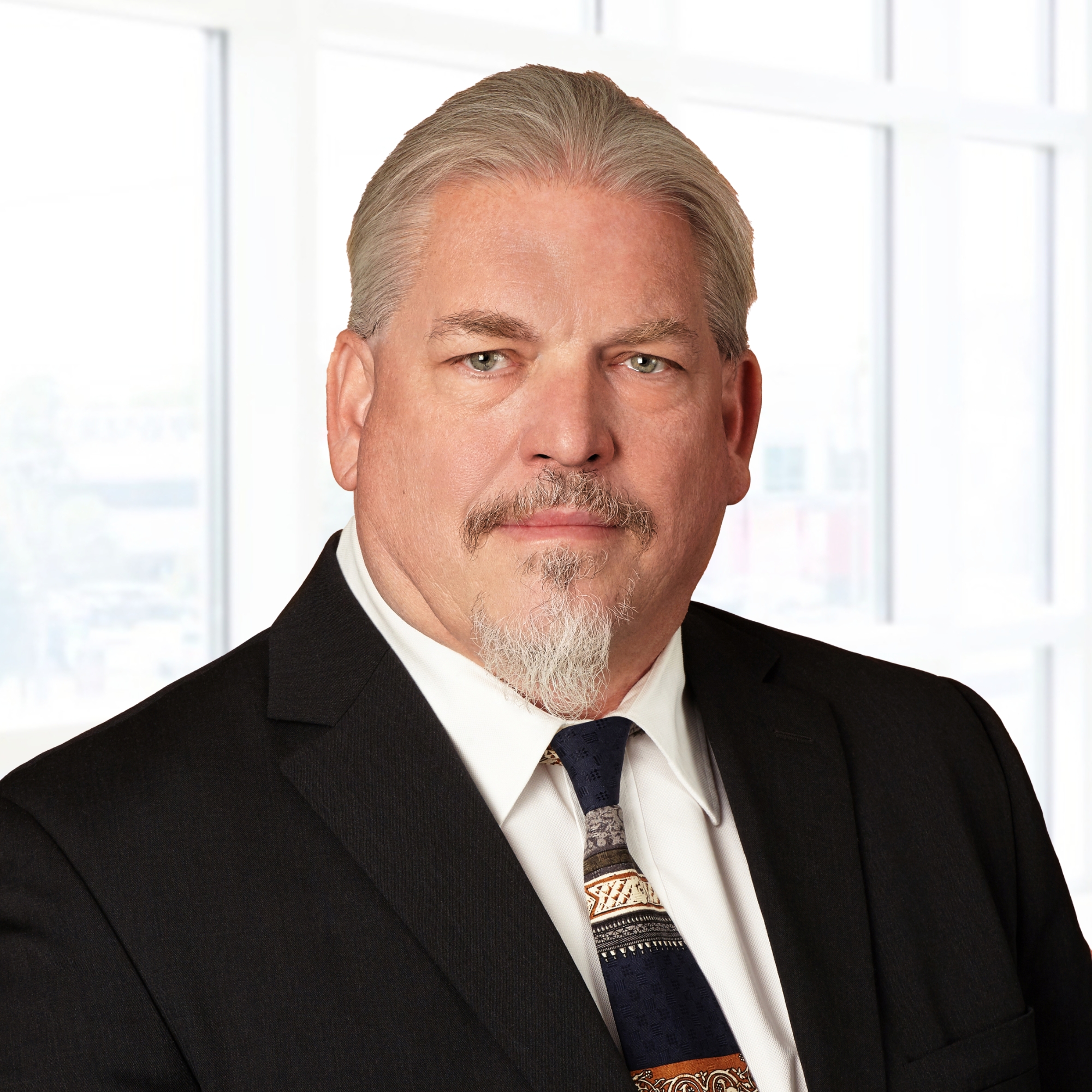 A man with gray hair and a goatee wearing a dark suit and patterned tie stands in front of a bright window.