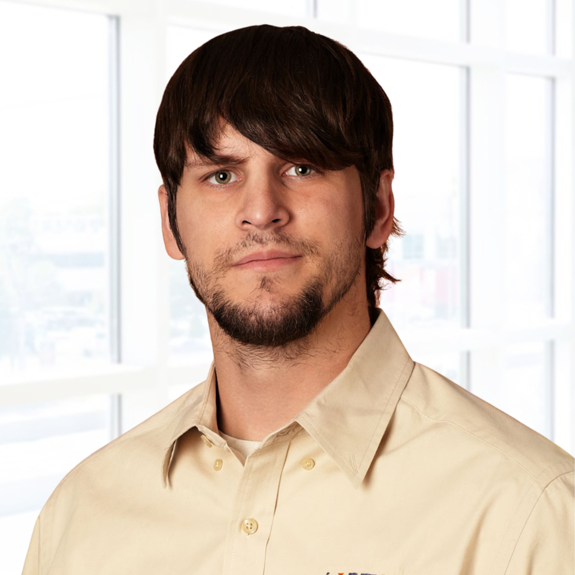 Person with dark hair and a beard wearing a beige collared shirt, standing in front of large windows with a blurred view outside.