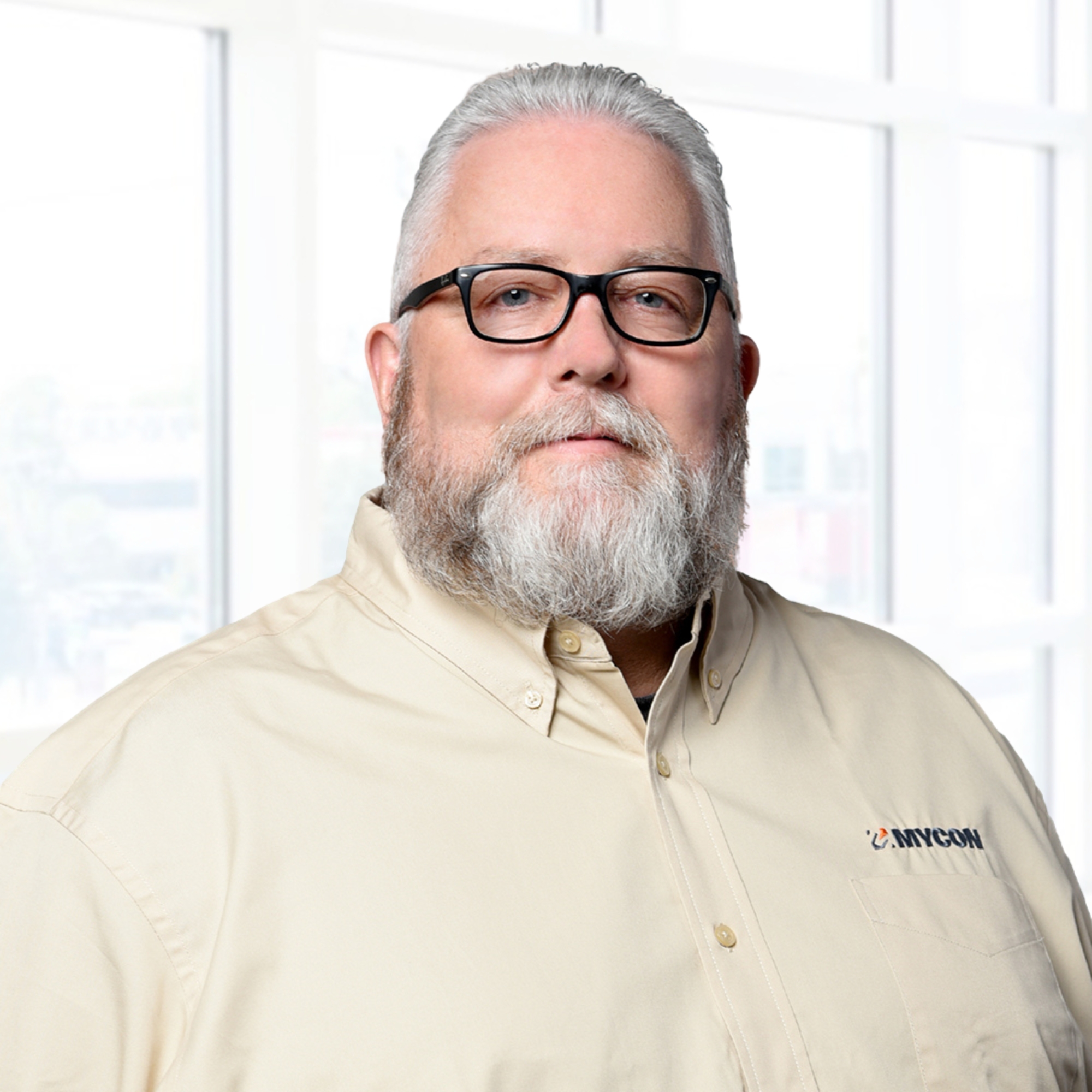 Man with glasses and a beard, wearing a beige shirt with "MYCOM" logo, stands in front of a bright window.