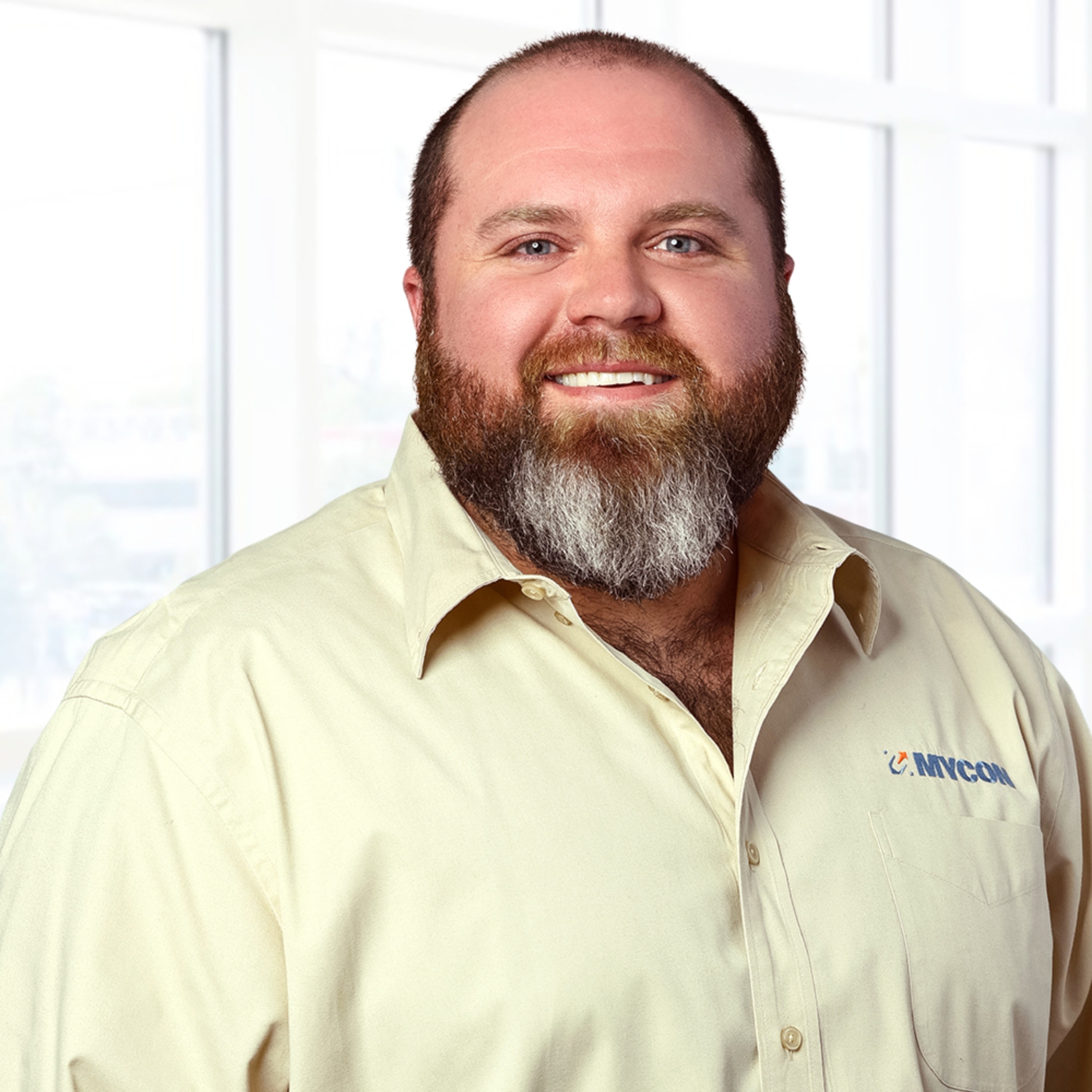 A bearded man in a light yellow shirt smiles in front of a bright window.