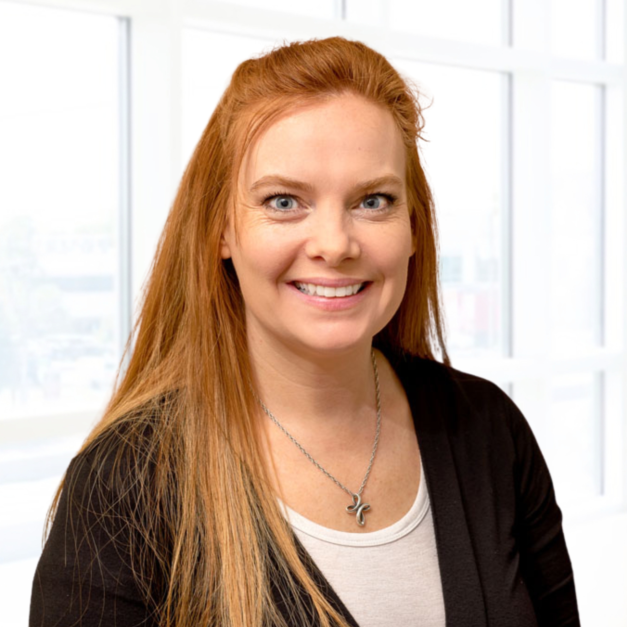 A woman with long red hair wearing a black cardigan and white shirt smiles in a bright room with large windows.