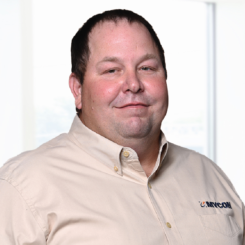 A man wearing a beige button-up shirt with a company logo stands in front of a light background. He has short dark hair and a slight smile.