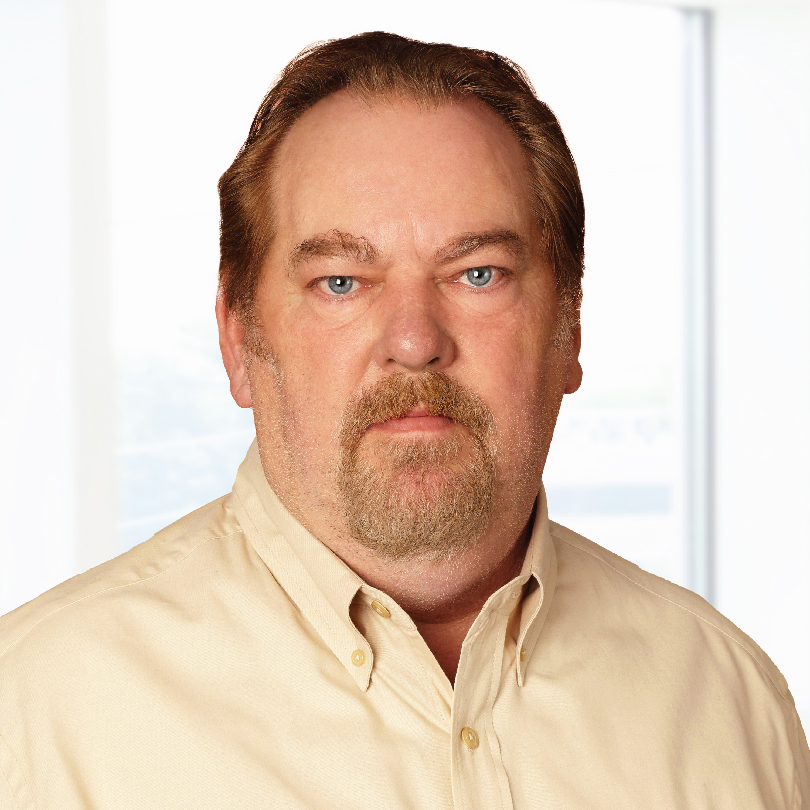 A middle-aged man with short, brown hair and a beard wearing a beige button-up shirt stands in front of a light background.
