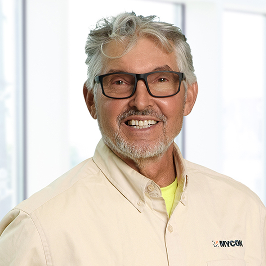 Smiling senior man with gray hair and glasses, wearing a beige shirt labeled "unicom," standing in a bright office near windows.