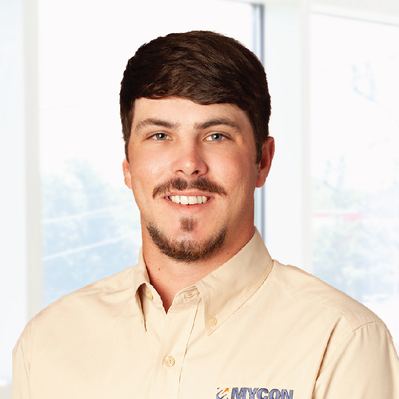Man with short brown hair and a beard, wearing a beige button-up shirt with the logo "MYCON" embroidered on it, standing against a bright background with large windows.