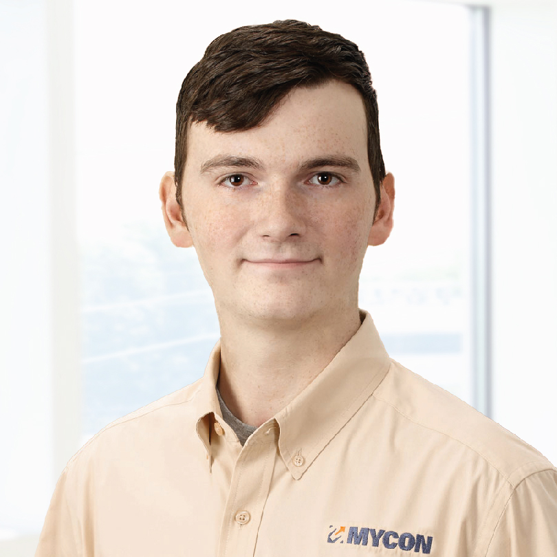 A person wearing a beige shirt with a "MYCON" logo stands in front of a bright, blurred background.