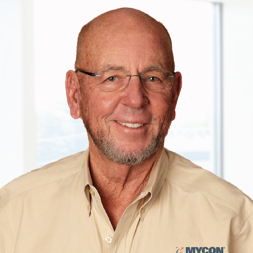 A bald man with glasses, wearing a beige shirt with a MYCON logo, stands smiling in a well-lit room.