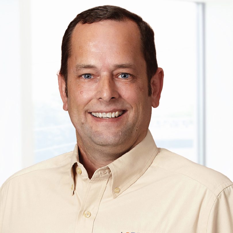A person with short brown hair and a beige collared shirt smiles against a light background.