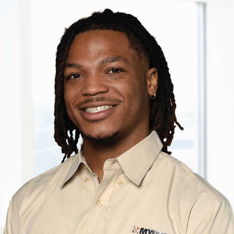 A person with long dreadlocks, wearing a beige collared shirt with a logo, smiling at the camera against a bright indoor backdrop.