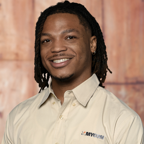 A man with dreadlocks smiling in front of a wooden wall.