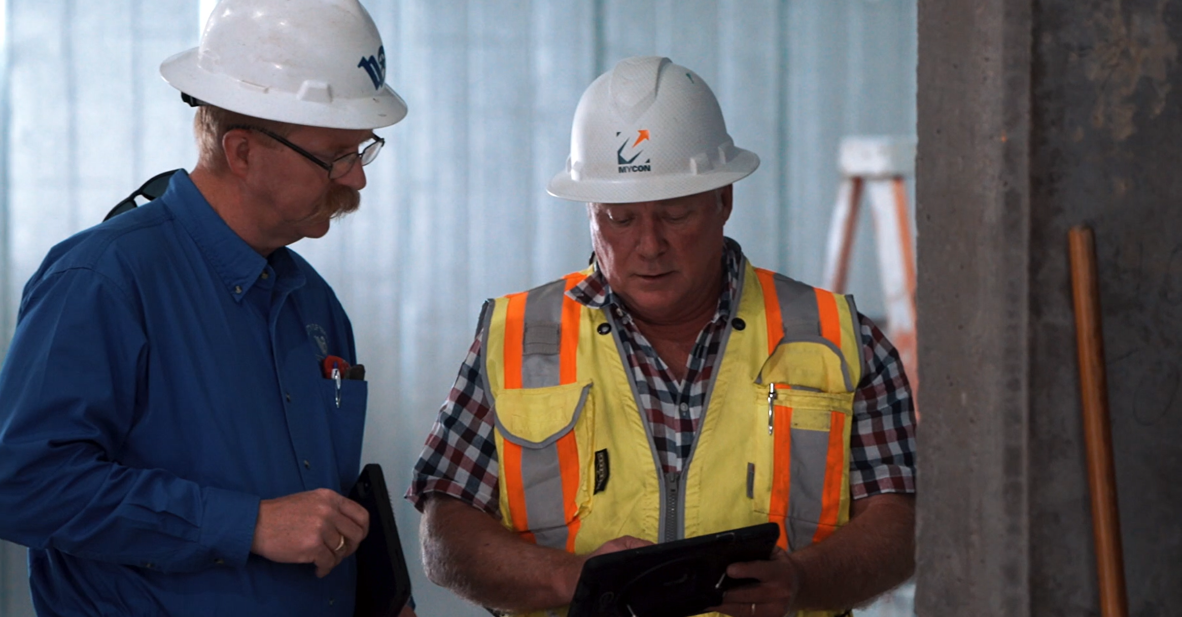 Two construction workers in hard hats looking at a tablet.