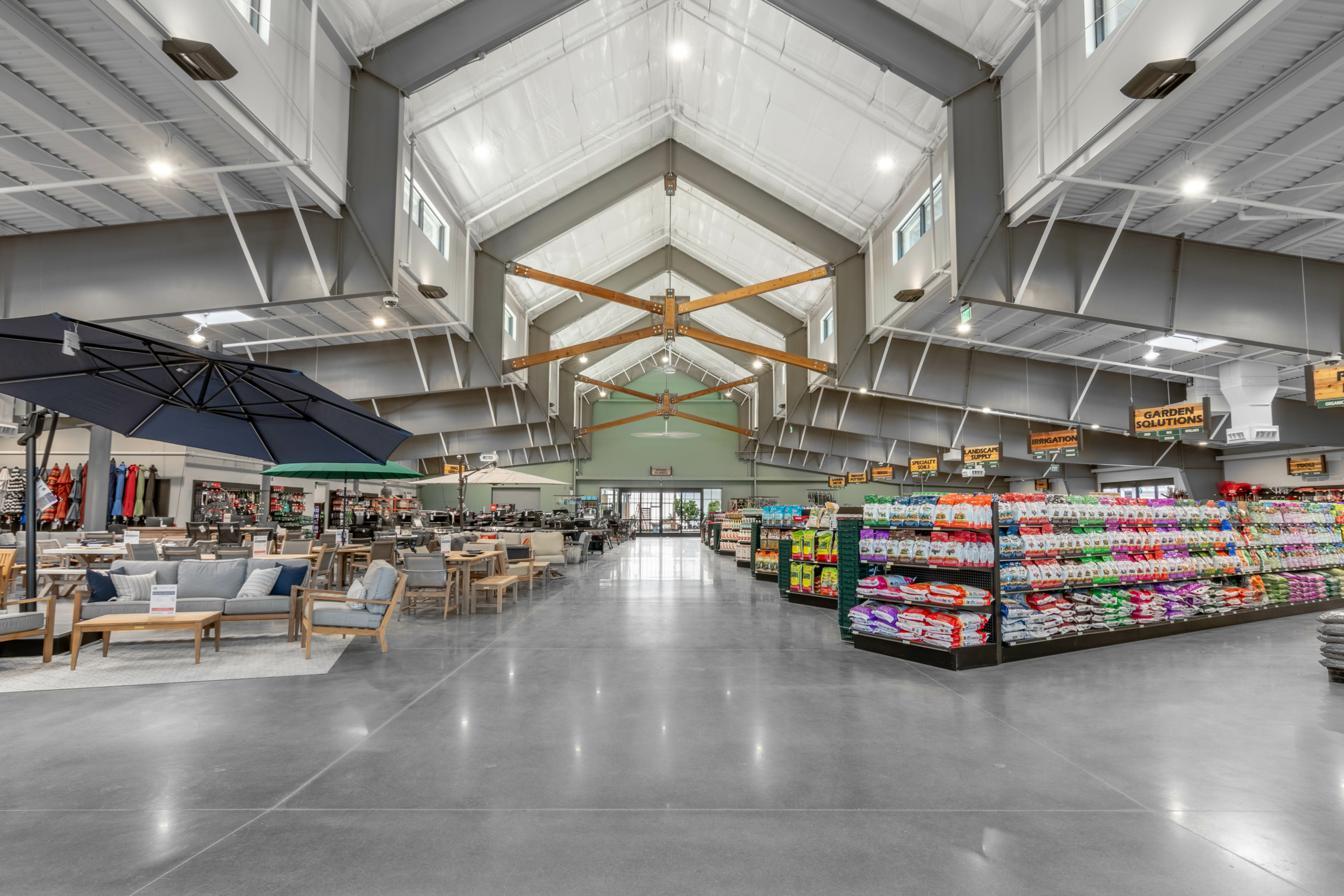 A spacious, well-lit store interior featuring outdoor furniture on the left and shelves stocked with various colorful products on the right. High ceilings and exposed beams are visible.