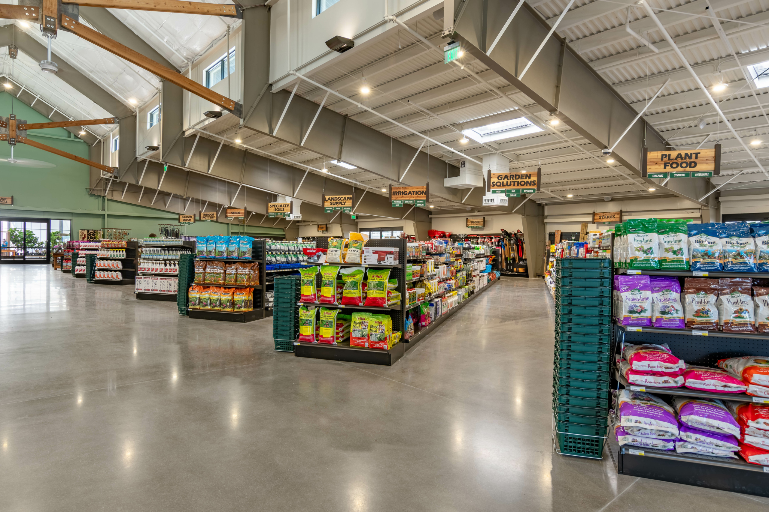 Interior view of a spacious, well-lit store with organized aisles displaying various gardening supplies and pet food, indicating clearly labeled sections like "Plant Food," "Garden Solutions," and "Wild Bird.