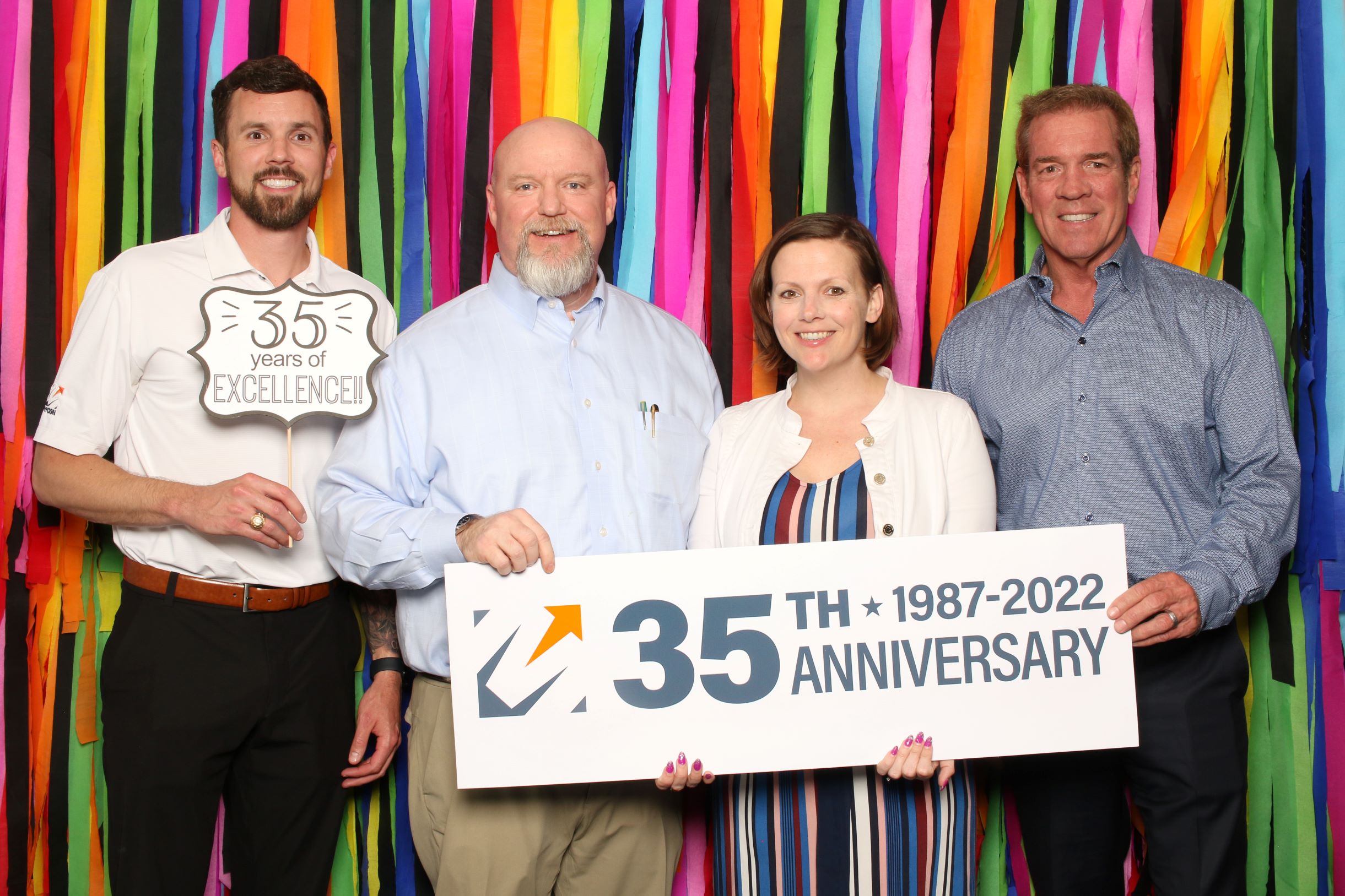 Four individuals posing with a sign celebrating a 35th anniversary, standing in front of a colorful striped background.