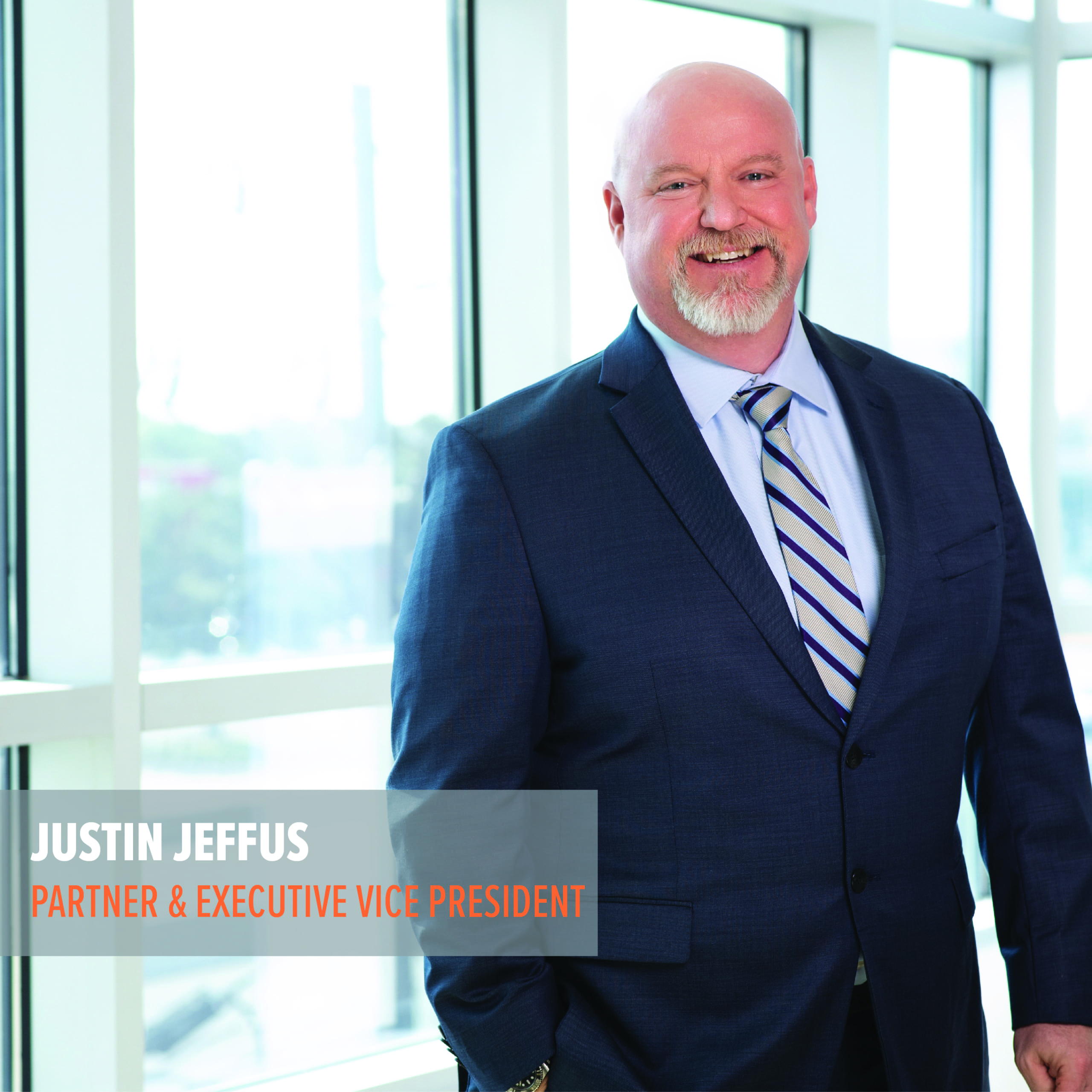 A professional portrait of a smiling executive man standing by the window, dressed in a suit with his name and title displayed as "justin jeffus, partner & executive vice president.