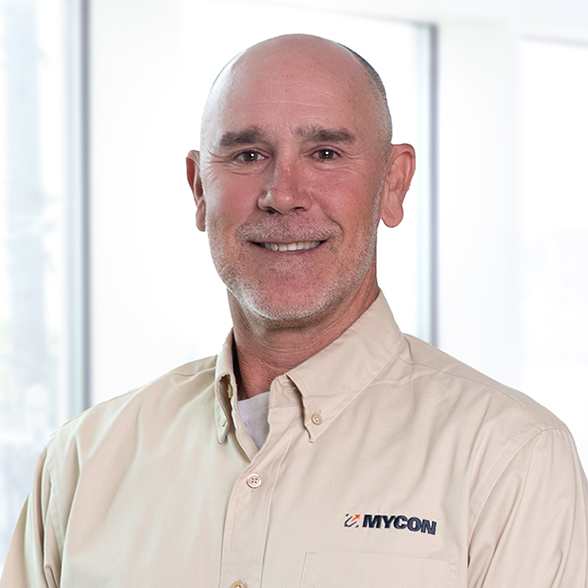 A smiling middle-aged man in a beige work shirt with "nycon" embroidered on it, standing in an office environment with large windows in the background.