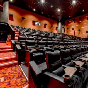 Interior of a modern cinema with rows of black leather recliner seats, red carpeting, and dim lighting. The image shows steps leading up to multiple rows of seats.