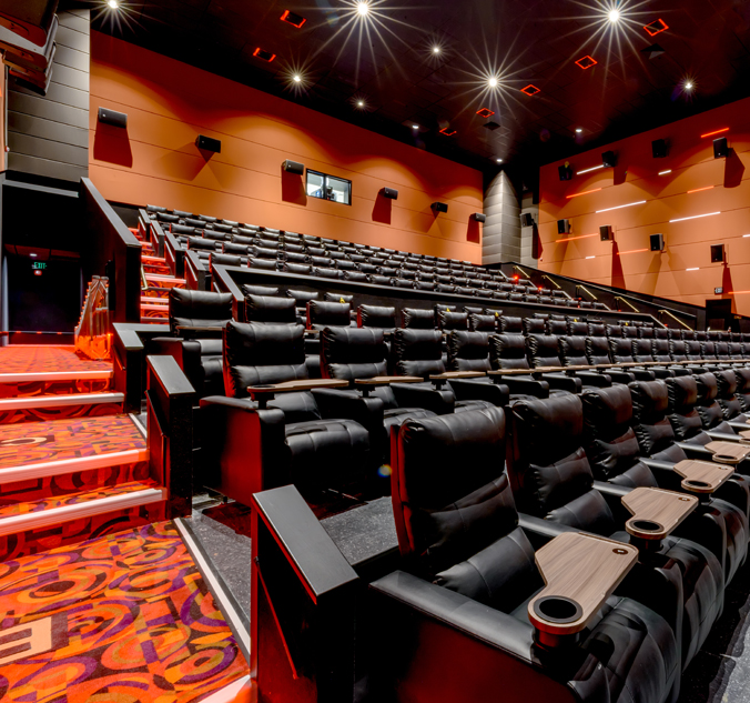 Interior of a modern cinema with rows of black leather recliner seats, red carpeting, and dim lighting. The image shows steps leading up to multiple rows of seats.
