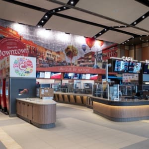 A modern movie theater concession stand with soft drink dispensers, snack displays, and a digital menu, featuring a mural of a historic downtown scene in the background.