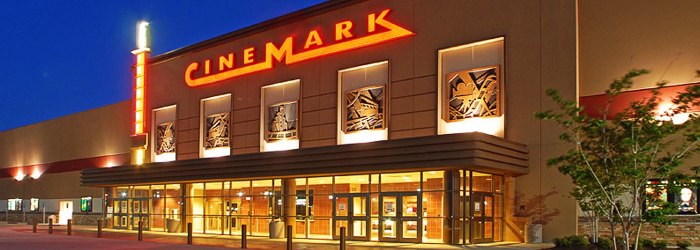 Front view of a Cinemark movie theater at night, illuminated by bright lights with large windows showcasing movie posters.