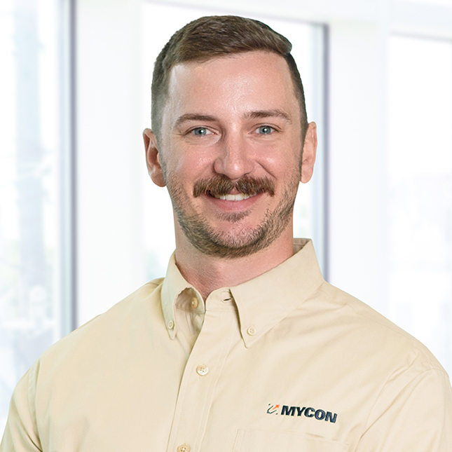 A man with a mustache smiling, wearing a light beige shirt with "nycon" logo, standing in front of a windowed backdrop.