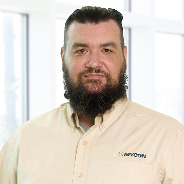 A man with a beard wearing a light beige shirt with a "mycon" logo, standing in front of a windowed office background.