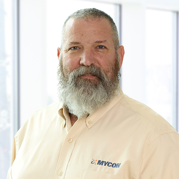 A bearded middle-aged man with a confident smile, wearing a beige shirt with a name logo, standing in an office environment.