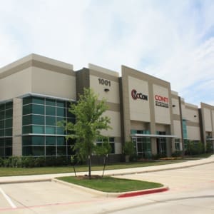 Exterior view of a modern office building with the address "1001" and logos for McCann Systems and Conti Systems on the front. There is a tree and landscaping in front of the building.