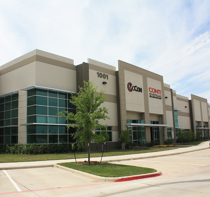 Exterior view of a modern office building with the address "1001" and logos for McCann Systems and Conti Systems on the front. There is a tree and landscaping in front of the building.