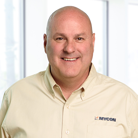 Smiling middle-aged man in a beige shirt with the logo "mycon" standing in a bright office environment.