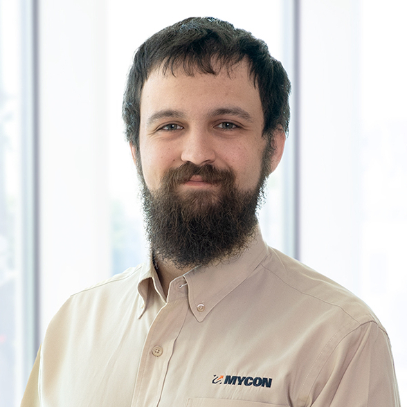 Man with a beard smiling, wearing a beige shirt with "envicom" logo, standing indoors with a window backdrop.