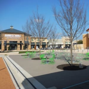 Outdoor plaza with green tables and chairs, leafless trees, and a Barnes & Noble bookstore in the background. Other retail stores are visible lining the walkway. Sky is clear and blue.