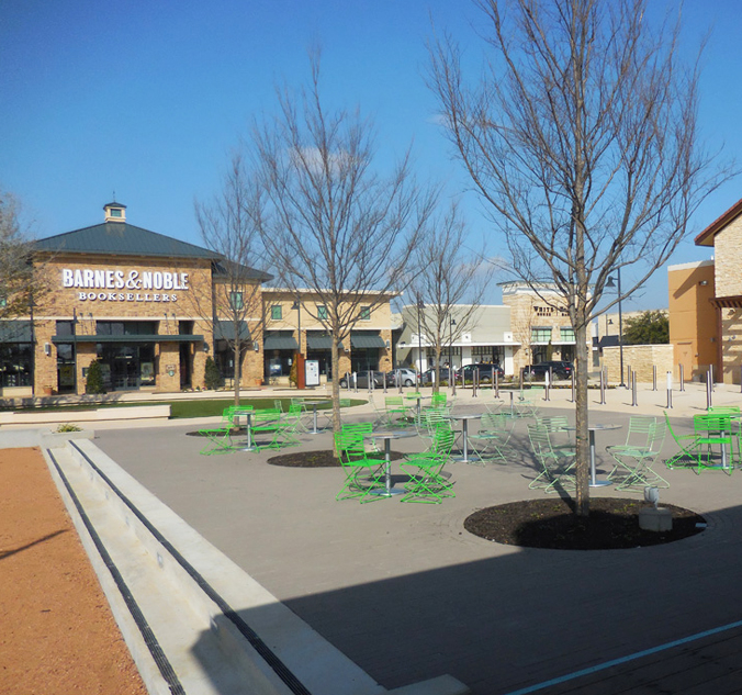 Outdoor plaza with green tables and chairs, leafless trees, and a Barnes & Noble bookstore in the background. Other retail stores are visible lining the walkway. Sky is clear and blue.