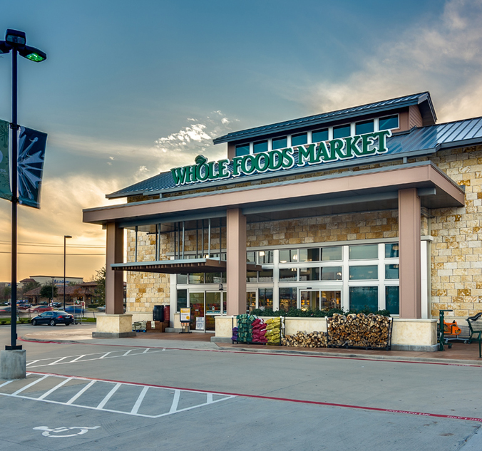 Whole Foods Market storefront with entrance under a large sign, surrounded by a parking lot with designated crossing areas and landscaping.