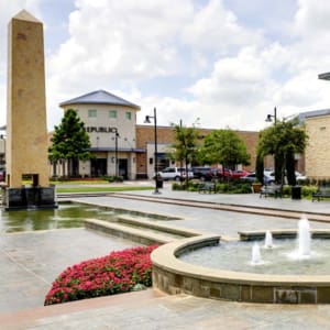 Outdoor plaza with an obelisk, fountains, seating areas, flower beds, and retail stores in the background on a sunny day.