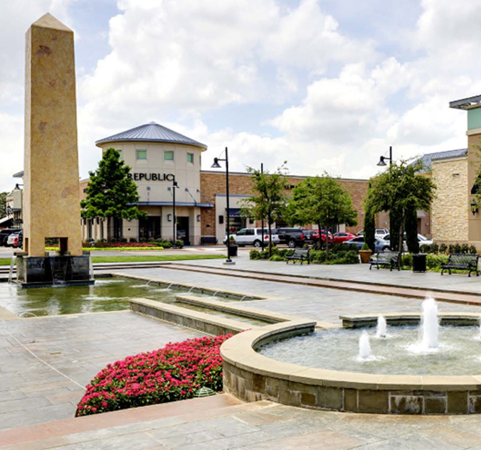Outdoor plaza with an obelisk, fountains, seating areas, flower beds, and retail stores in the background on a sunny day.
