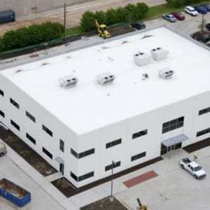 Aerial view of a two-story white rectangular building with a flat roof, rooftop HVAC units, and large parking area. Construction equipment and several cars are visible around the building.
