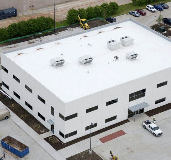 Aerial view of a two-story white rectangular building with a flat roof, rooftop HVAC units, and large parking area. Construction equipment and several cars are visible around the building.