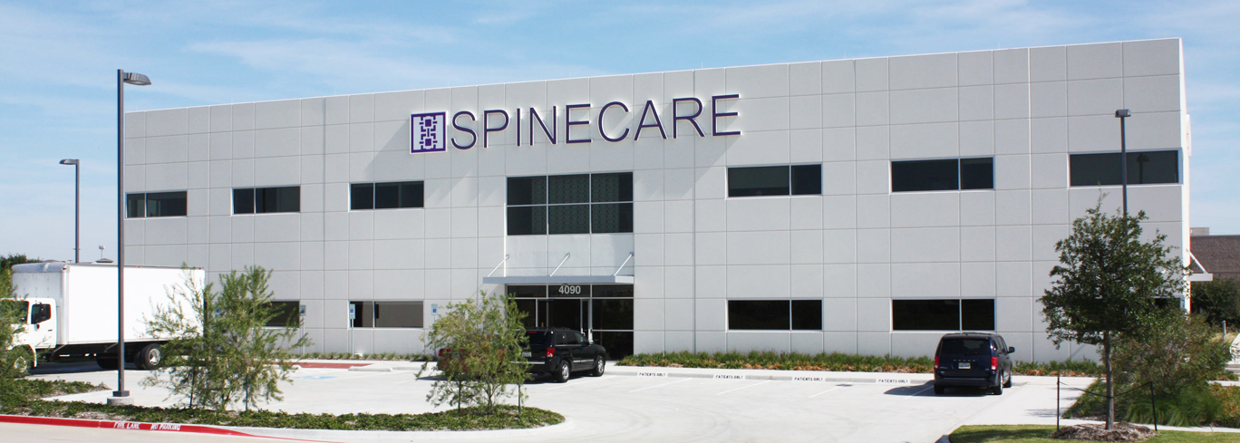 A two-story white commercial building with the sign "SPINECARE" and a logo on the front. The entrance has a covered doorway, and there are a few cars and a truck in the parking lot.