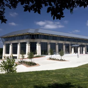 A large building with a modern, metal roof design, stone exterior walls, and ample windows is situated among landscaped greenery under a clear blue sky.