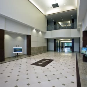 Spacious modern lobby with high ceilings, tiled floor, a painting on an easel, and framed pictures. Large windows in the background provide natural light. Balcony overlooks the lobby area.